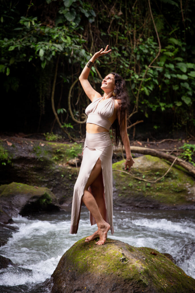 Amira Attar standing on a rock by a jungle waterfall, one arm raised